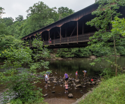 Children play in shallow river water below a covered bridge