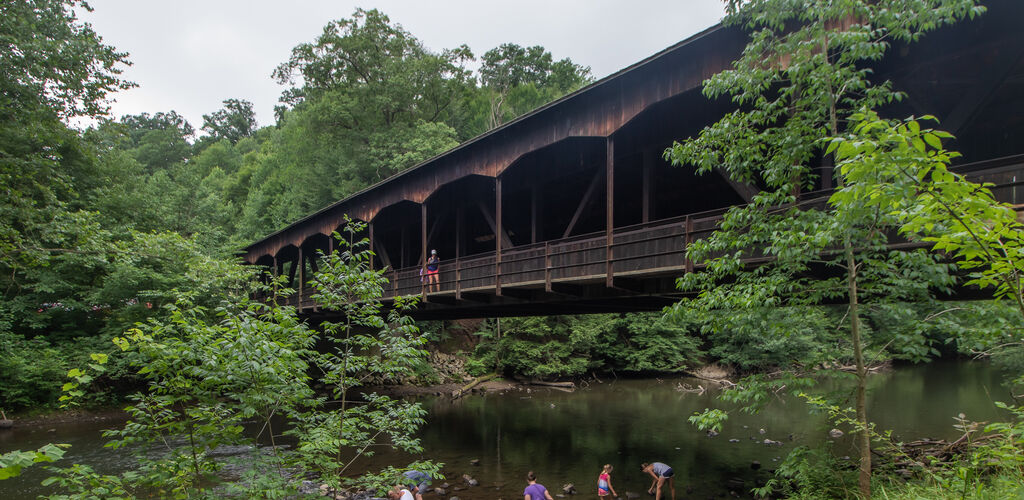 Children play in shallow river water below a covered bridge